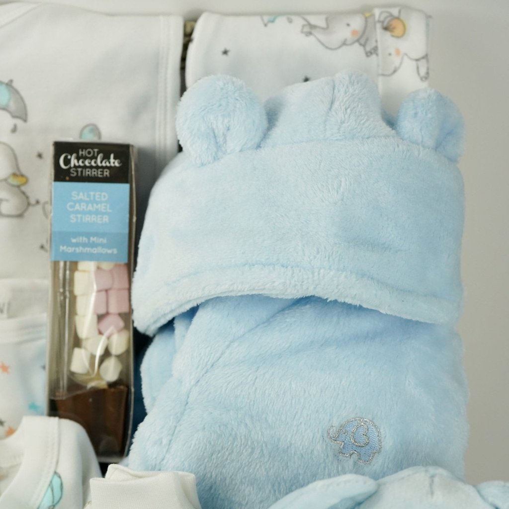 baby boy hamper gift includes white baby layette with grey elephants with blue accents, blue baby dressing gown, blue elephant soft, blue knit booties, chocolate stirring spoon, baby taggie comfort blanket