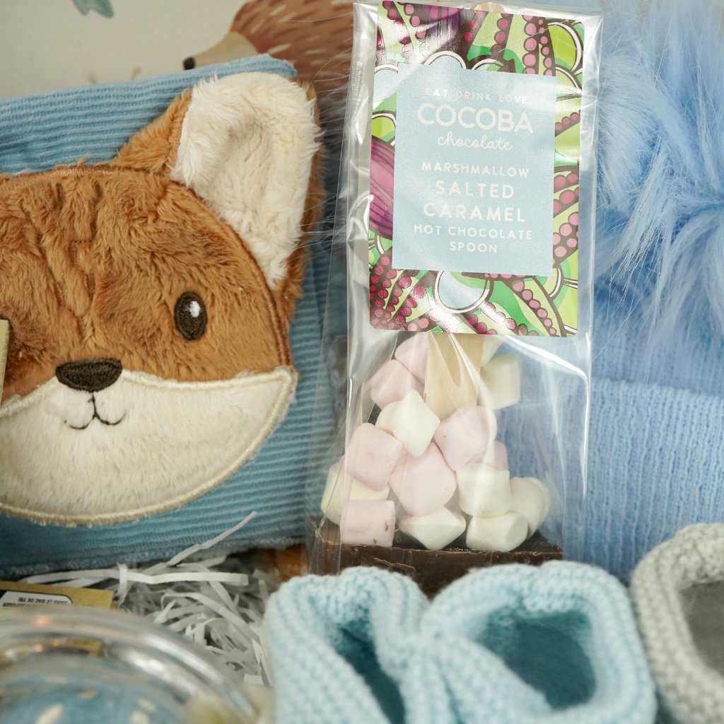 cardboard suitcase with soft activity book in blue with fox head design, fox soft rattle, chocolate spoon with mallows, blue fluffy pompom hat, two pairs of booties 