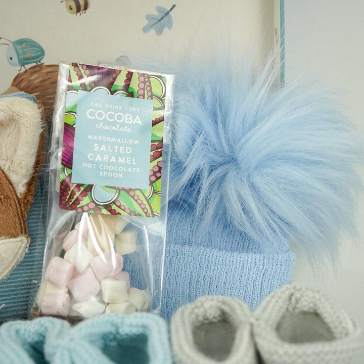 cardboard suitcase with soft activity book in blue with fox head design, fox soft rattle, chocolate spoon with mallows, blue fluffy pompom hat, two pairs of booties 