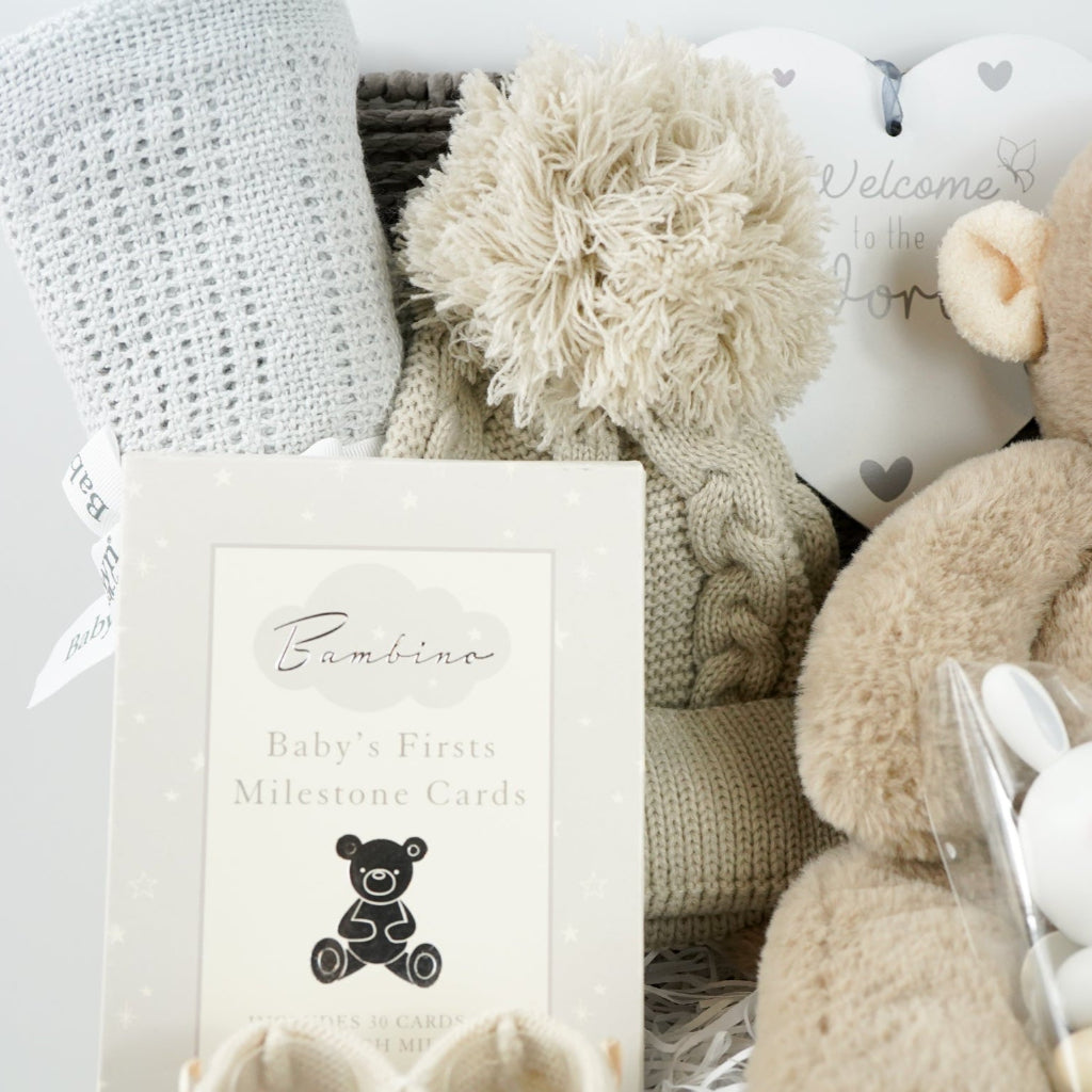 Baby grey hamper basket with grey cellular baby blanket, baby biscuit coloured pom pom hat, heart hanging nursery plaque, Baby milestone Cards, baby booties in biscuit colour, wooden stacking toy and soft eco friendly baby soft toy