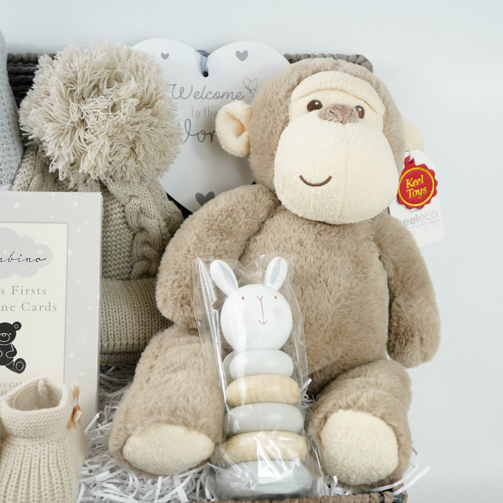 Baby grey hamper basket with grey cellular baby blanket, baby biscuit coloured pom pom hat, heart hanging nursery plaque, Baby milestone Cards, baby booties in biscuit colour, wooden stacking toy and soft eco friendly baby soft toy