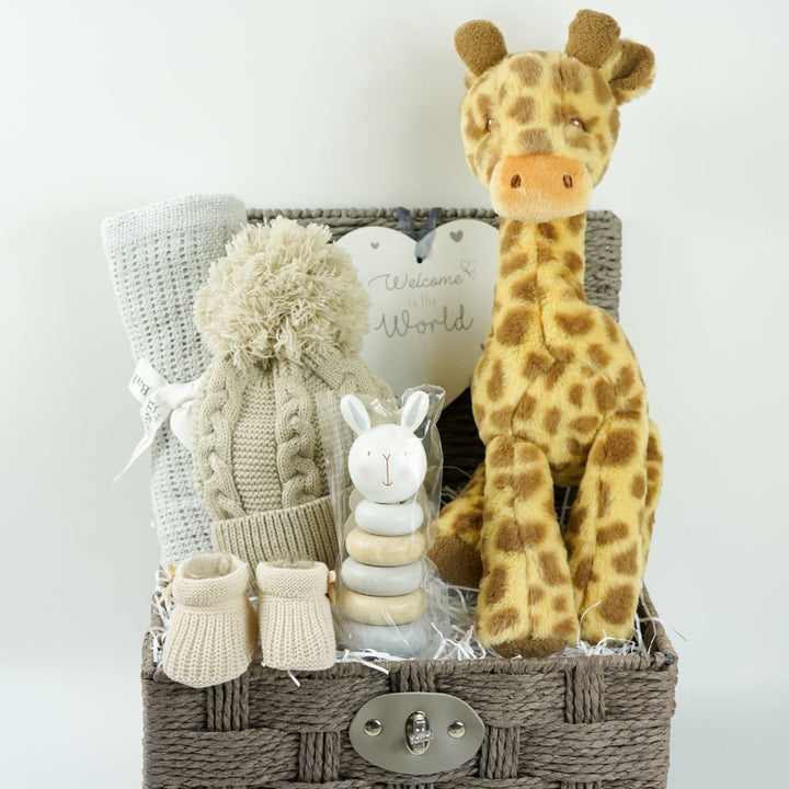 Grey hamper basket with baby gifts including grey cellular blanket, biscuit coloured knit pompom hat, wooden stacking toy, nursery plaque in heart shape with 'welcome to the world', soft giraffe