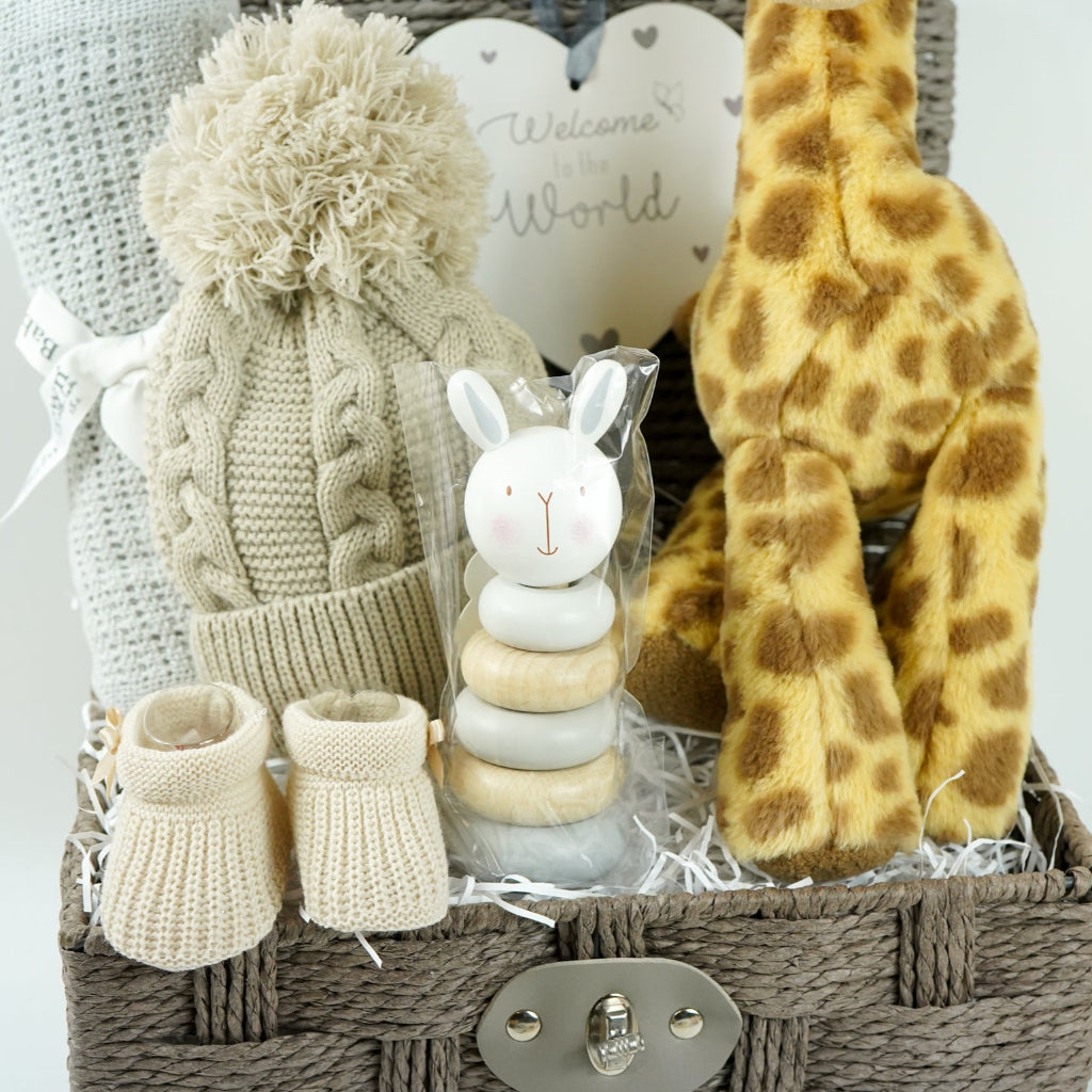 Grey hamper basket with baby gifts including grey cellular blanket, biscuit coloured knit pompom hat, wooden stacking toy, nursery plaque in heart shape with 'welcome to the world', soft giraffe