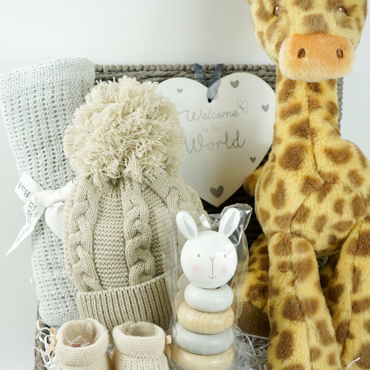 Grey hamper basket with baby gifts including grey cellular blanket, biscuit coloured knit pompom hat, wooden stacking toy, nursery plaque in heart shape with 'welcome to the world', soft giraffe