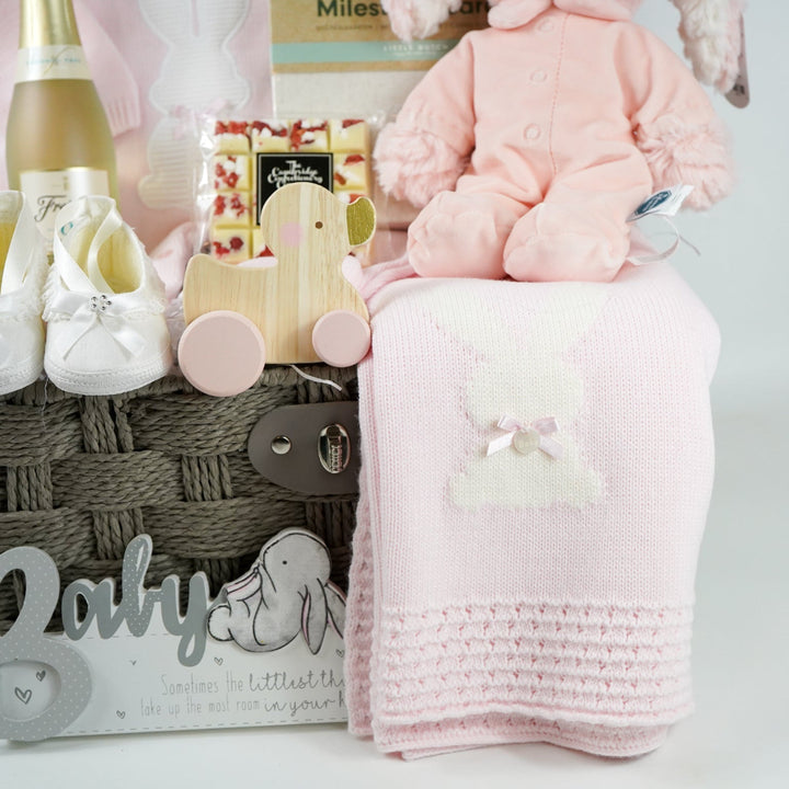 baby girl hamper basket includes fine knit pink romper with feet and white bunny  and matching blanket, small bottle alcohol free sparkling wine, satin baby shoes, white chocolate eton mess bar, wooden push along duck, pink bunny in velour romper, memorable moments cars in a box, baby plaque