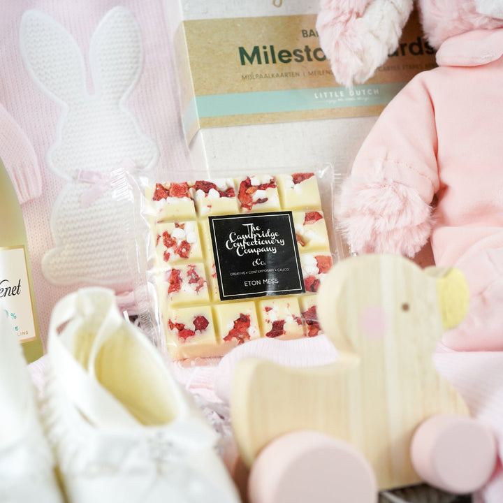 baby girl hamper basket includes fine knit pink romper with feet and white bunny  and matching blanket, small bottle alcohol free sparkling wine, satin baby shoes, white chocolate eton mess bar, wooden push along duck, pink bunny in velour romper, memorable moments cars in a box, baby plaque