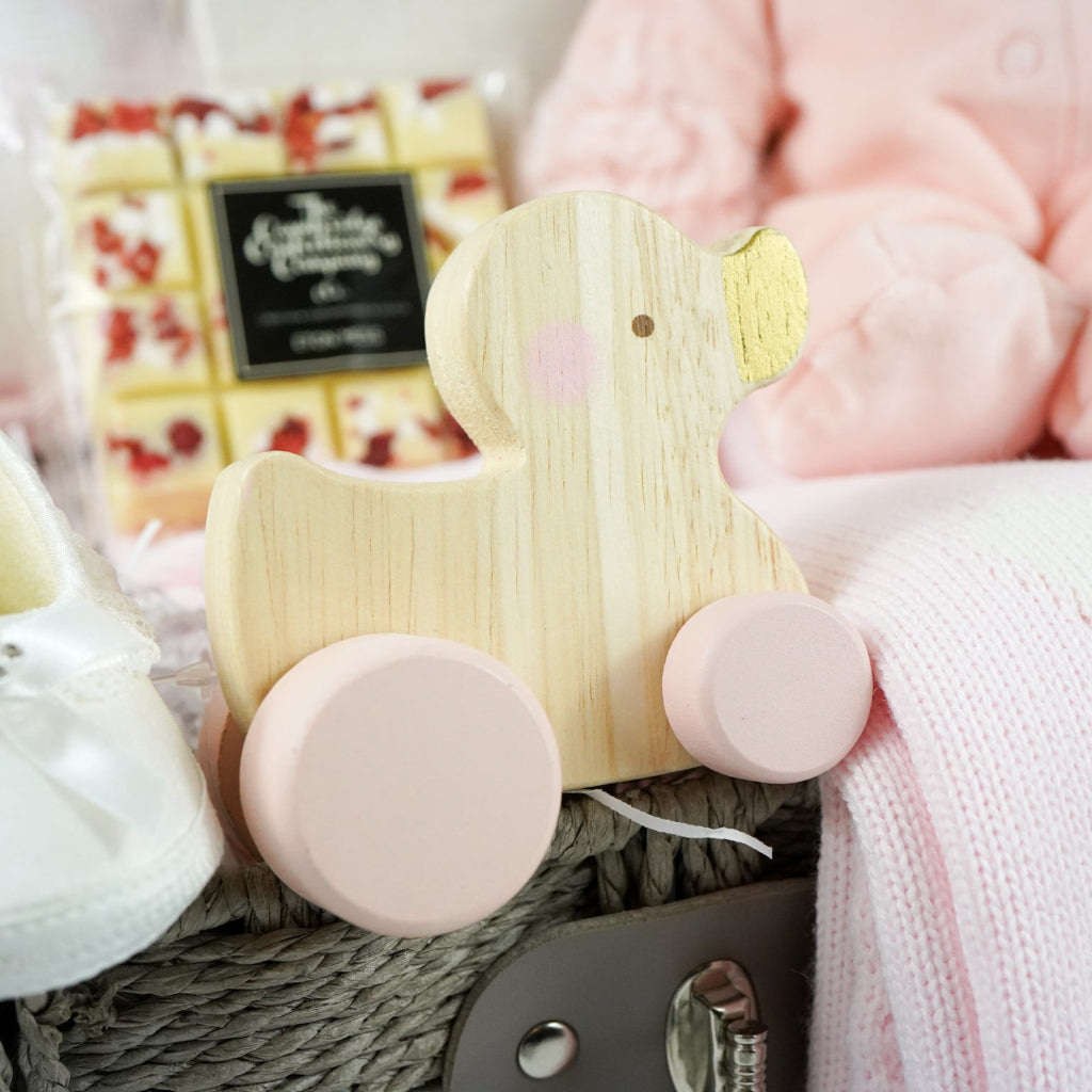 baby girl hamper basket includes fine knit pink romper with feet and white bunny  and matching blanket, small bottle alcohol free sparkling wine, satin baby shoes, white chocolate eton mess bar, wooden push along duck, pink bunny in velour romper, memorable moments cars in a box, baby plaque