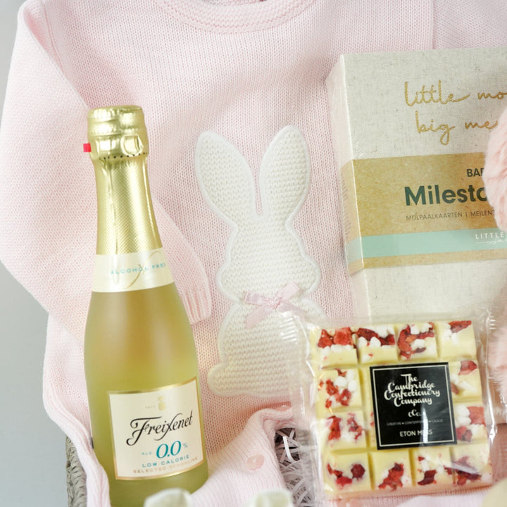 baby girl hamper basket includes fine knit pink romper with feet and white bunny  and matching blanket, small bottle alcohol free sparkling wine, satin baby shoes, white chocolate eton mess bar, wooden push along duck, pink bunny in velour romper, memorable moments cars in a box, baby plaque