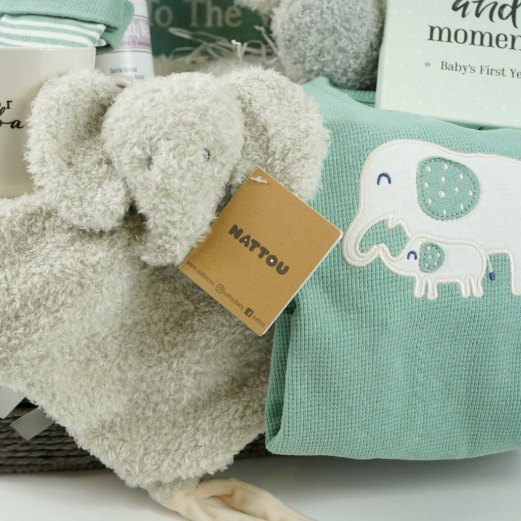 Baby hamper basket with soft grey fluffy elephant by Nattou and matching baby comforter, softt organic baby blanket in green and cream, organic green waffle effect baby romper with cream elephant design, green baby socks with elephants, knit beanie hat in green with elephants, baby porcelain mug with gold writing for baby, organic baby wash,  Welcome o the world baby book