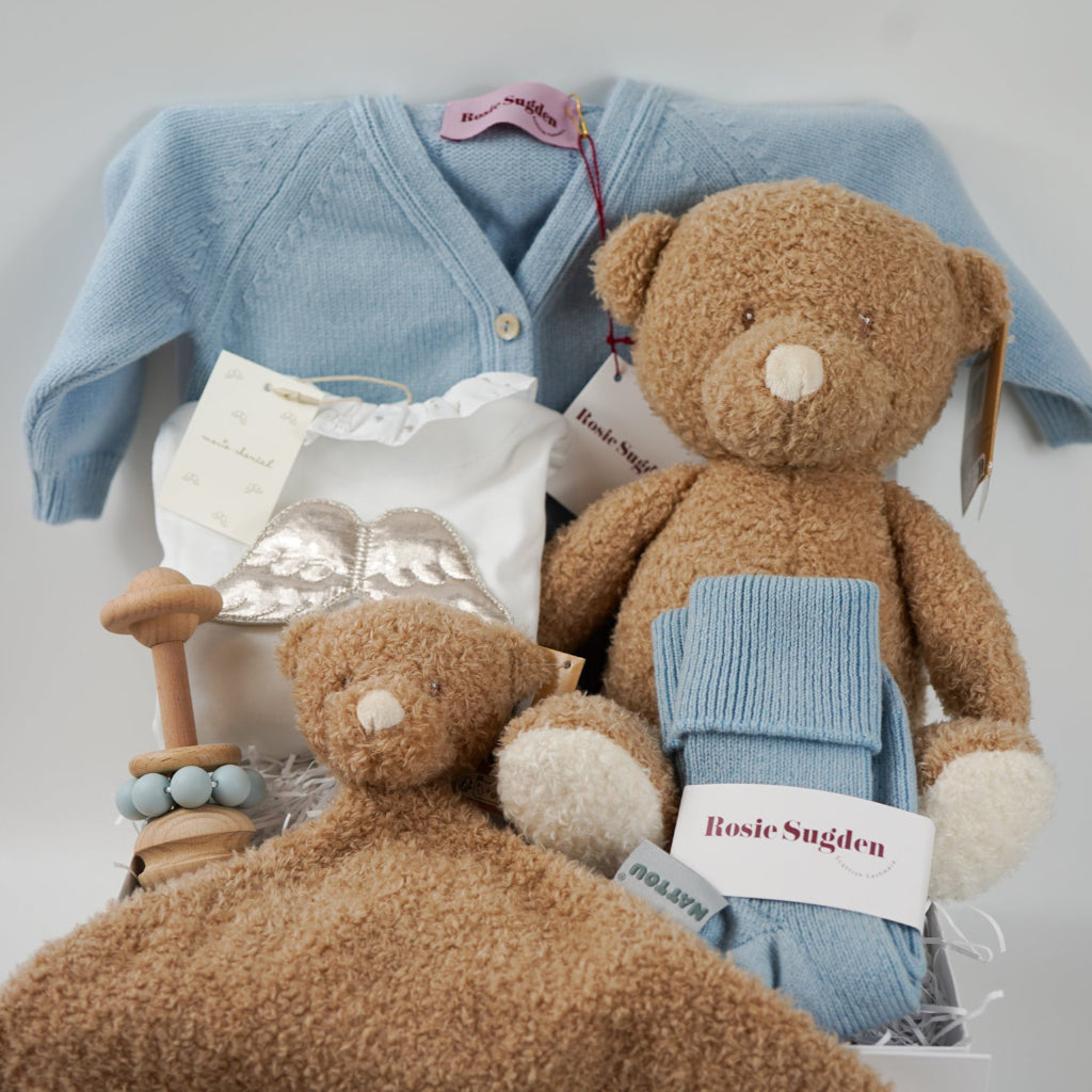 Luxury baby gift hamper box with pale blue cashmere baby cardigan, white luxury baby sleepsuit with frilled collar and silver angel wings on the back, wooden baby rattle with blue silicone teething beads, matching cashmere socks for mum in pal blue, soft teddy and matching baby comforter 