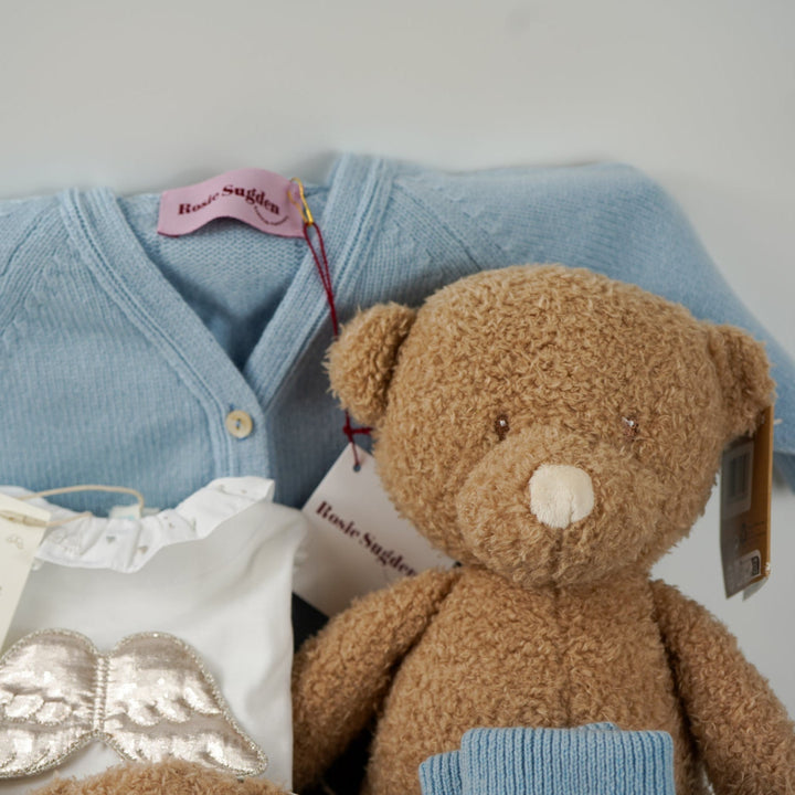 Luxury baby gift hamper box with pale blue cashmere baby cardigan, white luxury baby sleepsuit with frilled collar and silver angel wings on the back, wooden baby rattle with blue silicone teething beads, matching cashmere socks for mum in pal blue, soft teddy and matching baby comforter 