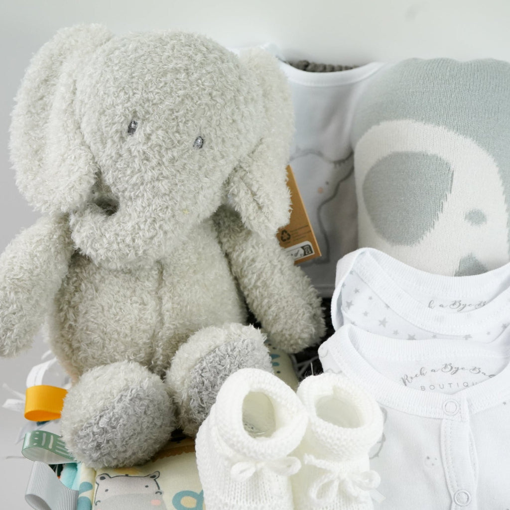 eutral baby hamper in grey rope basket, grey nattou soft elephant, grey and white soft baby blanket, taggie blanket with lion, elephants and zebras faces  with fleece backing, white baby layette set with grey elephant  design includes baby sleepsuit, baby bodysuit, hat, mittens and bib