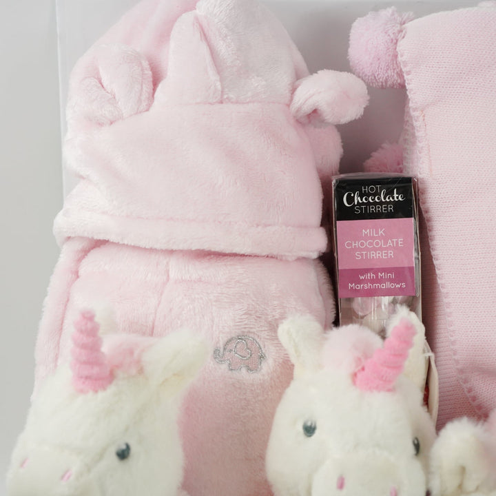 white hamper box with pink baby gown, soft eco friendly unicorn oft toy, unicorn rattle and baby comforter , pink and cream baby blanket with hearts and pom poms, chocolate stirrer spoon