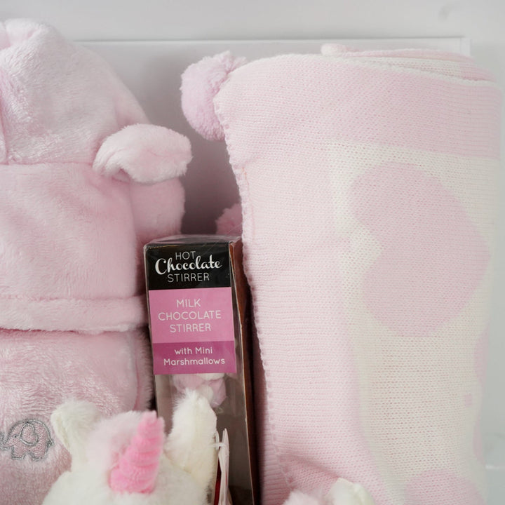 white hamper box with pink baby gown, soft eco friendly unicorn oft toy, unicorn rattle and baby comforter , pink and cream baby blanket with hearts and pom poms, chocolate stirrer spoon