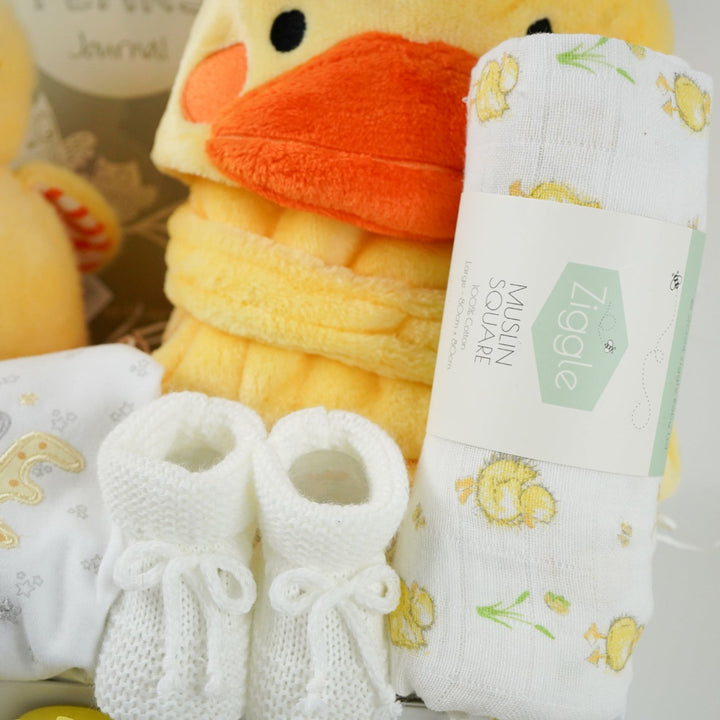 white hamper box with yellow baby dressing gown with duck face hood, soft cuddly yellow duck toy, white muslin square with yellow duck pattern., white knit baby booties, white baby sleep suit with zebra, giraffe and elephant and applique NEWBIE all in grey, yellow and orange, Early years baby journal 
