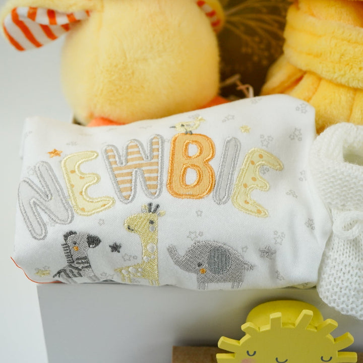 white hamper box with yellow baby dressing gown with duck face hood, soft cuddly yellow duck toy, white muslin square with yellow duck pattern., white knit baby booties, white baby sleep suit with zebra, giraffe and elephant and applique NEWBIE all in grey, yellow and orange, Early years baby journal 