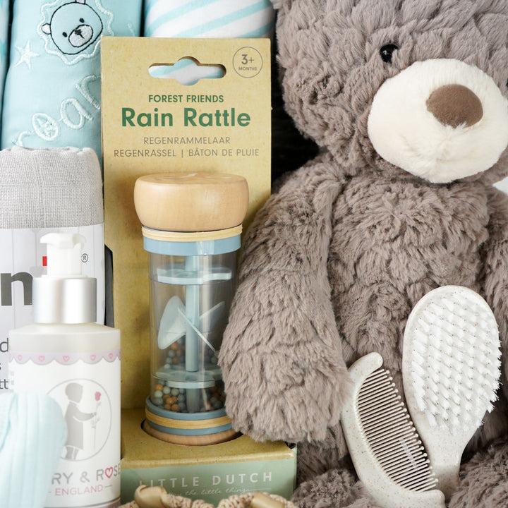grey paper rope hamper basket with three blue and white hooded towels, baby rain rattle, 2 muslins, 3 wash clothes 
organic baby bath, blue ribbed zip up baby sleepsuit, caramel bunny knit baby shoes, baby brush and comb, soft brown/grey baby teddy