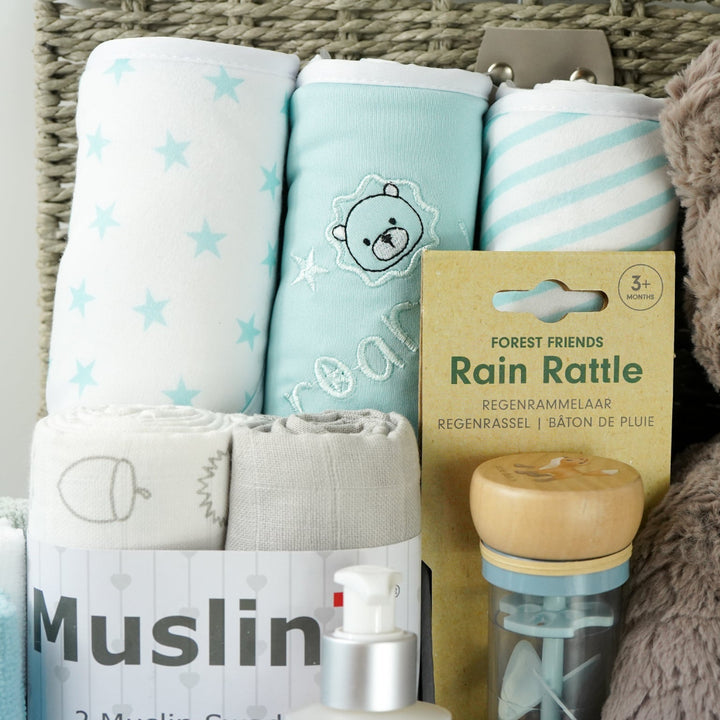 grey paper rope hamper basket with three blue and white hooded towels, baby rain rattle, 2 muslins, 3 wash clothes 
organic baby bath, blue ribbed zip up baby sleepsuit, caramel bunny knit baby shoes, baby brush and comb, soft brown/grey baby teddy