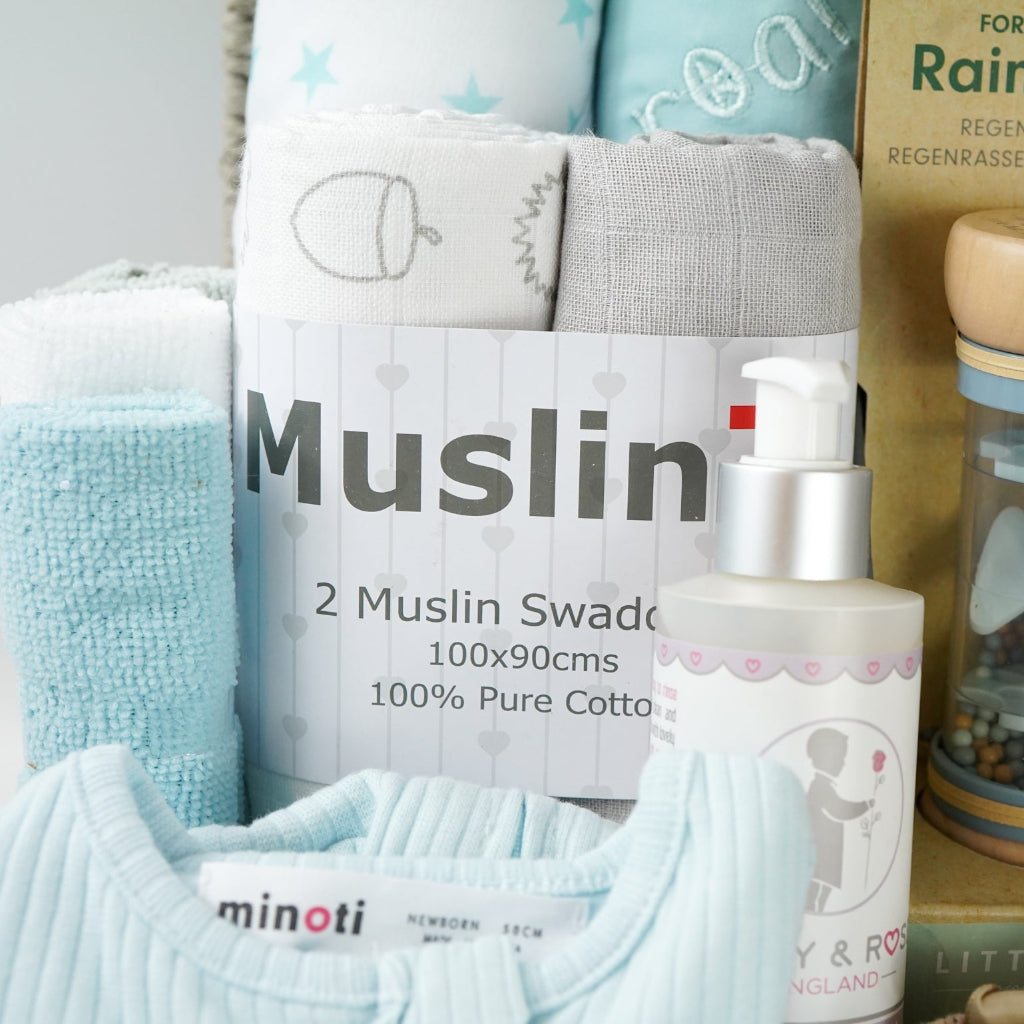 grey paper rope hamper basket with three blue and white hooded towels, baby rain rattle, 2 muslins, 3 wash clothes 
organic baby bath, blue ribbed zip up baby sleepsuit, caramel bunny knit baby shoes, baby brush and comb, soft brown/grey baby teddy