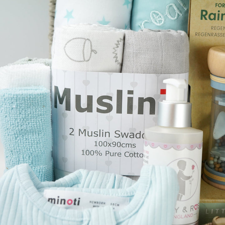 grey paper rope hamper basket with three blue and white hooded towels, baby rain rattle, 2 muslins, 3 wash clothes 
organic baby bath, blue ribbed zip up baby sleepsuit, caramel bunny knit baby shoes, baby brush and comb, soft brown/grey baby teddy