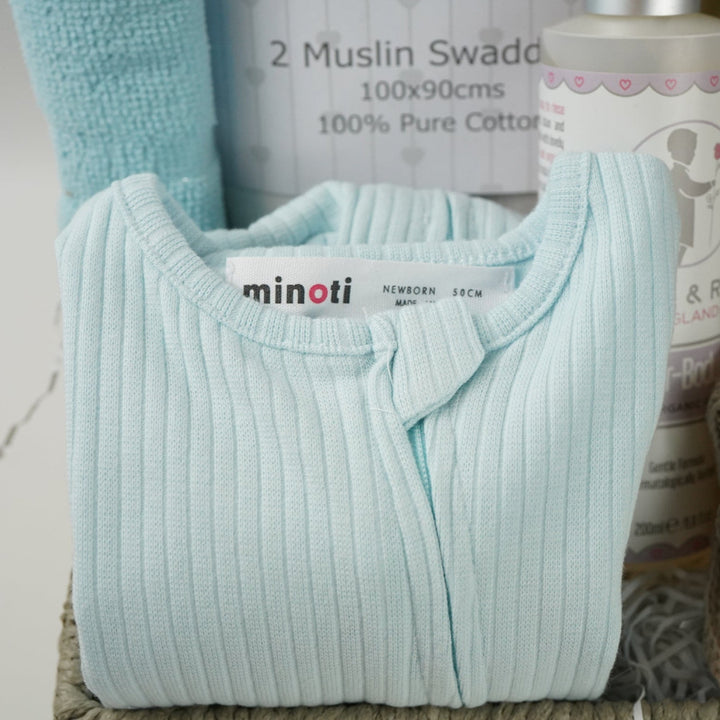 grey paper rope hamper basket with three blue and white hooded towels, baby rain rattle, 2 muslins, 3 wash clothes 
organic baby bath, blue ribbed zip up baby sleepsuit, caramel bunny knit baby shoes, baby brush and comb, soft brown/grey baby teddy