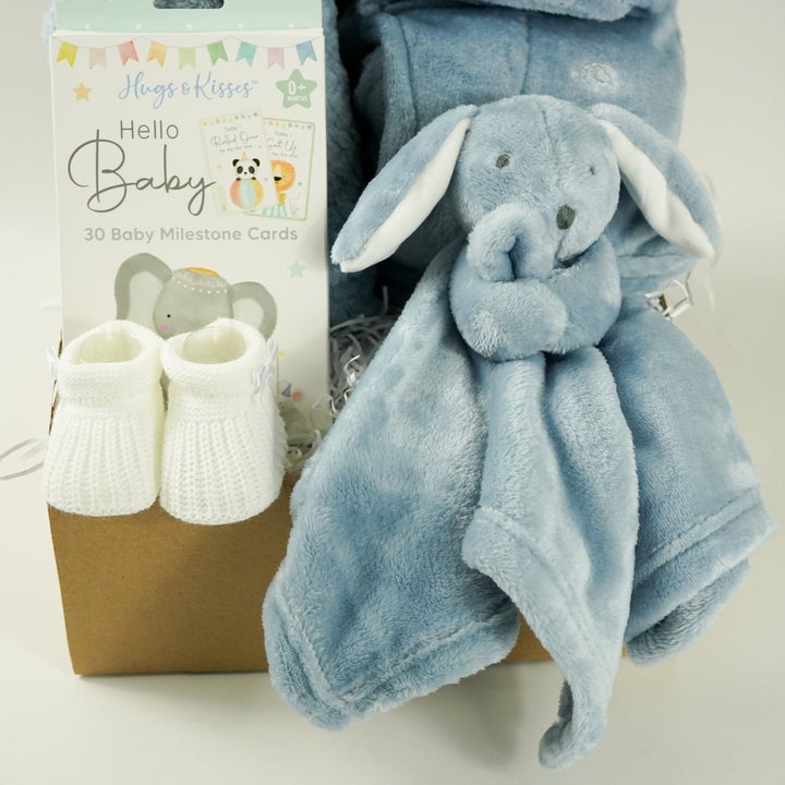 Baby gift set with blue blanket, booties, and comfort toy in a box on a light blue background