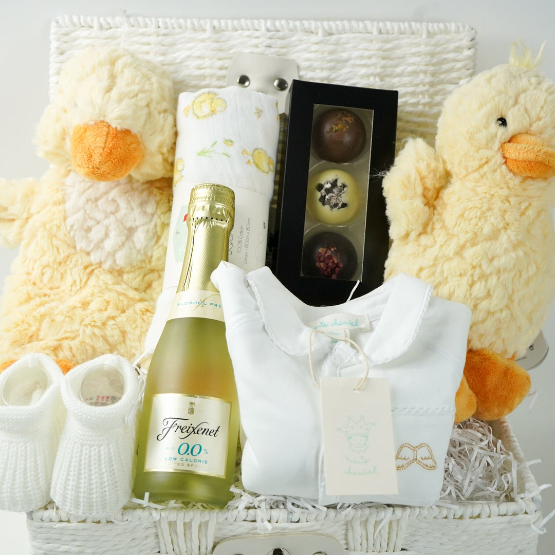 luxury baby gift hamper basket with yellow duck lovey and yellow duck , white muslin square with yellow ducks , white booties , luxury baby sleepsuit in white with gold angel wings embroidered on front  pocket and picot edging, alcohol free sparking wine and chocolates 