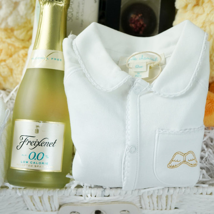 luxury baby gift hamper basket with yellow duck lovey and yellow duck , white muslin square with yellow ducks , white booties , luxury baby sleepsuit in white with gold angel wings embroidered on front  pocket and picot edging, alcohol free sparking wine and chocolates 
