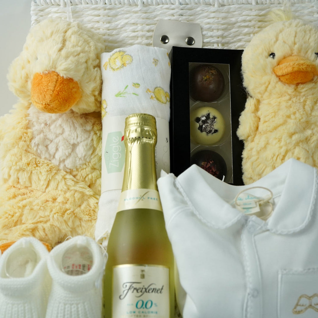 luxury baby gift hamper basket with yellow duck lovey and yellow duck , white muslin square with yellow ducks , white booties , luxury baby sleepsuit in white with gold angel wings embroidered on front  pocket and picot edging, alcohol free sparking wine and chocolates 
