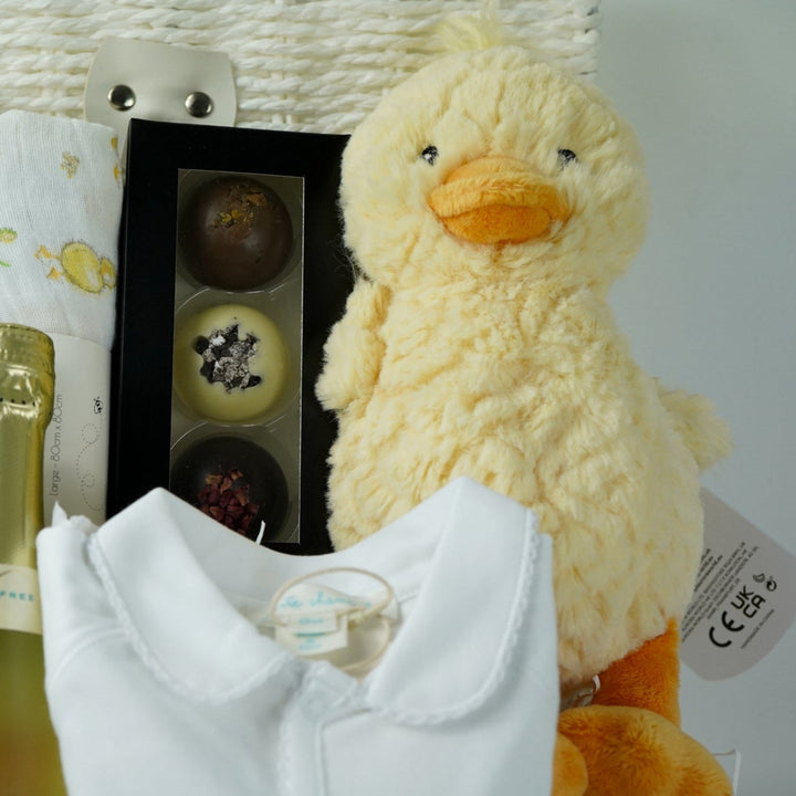 luxury baby gift hamper basket with yellow duck lovey and yellow duck , white muslin square with yellow ducks , white booties , luxury baby sleepsuit in white with gold angel wings embroidered on front  pocket and picot edging, alcohol free sparking wine and chocolates 