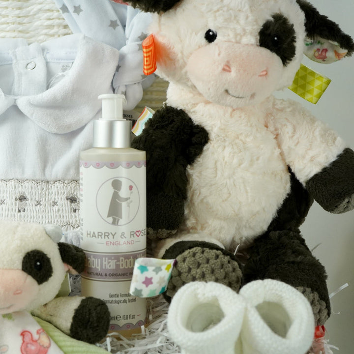 baby gift basket with a cow comforter and soft cow taggies toy, white baby sleepsuit with silver smocked sheep on bodice, organic toiletries, white knit booties and white with silver stars knot hat