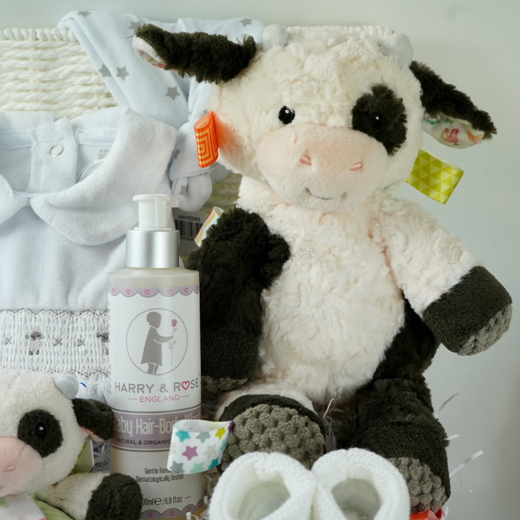 baby gift basket with a cow comforter and soft cow taggies toy, white baby sleepsuit with silver smocked sheep on bodice, organic toiletries, white knit booties and white with silver stars knot hat
