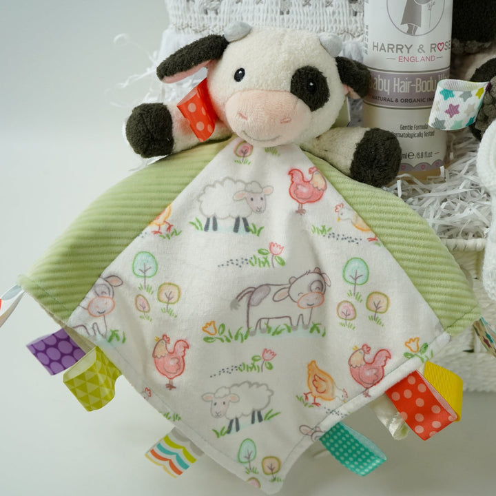 baby gift basket with a cow comforter and soft cow taggies toy, white baby sleepsuit with silver smocked sheep on bodice, organic toiletries, white knit booties and white with silver stars knot hat