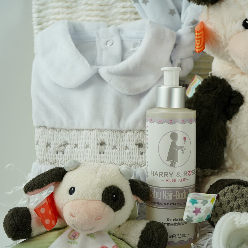 baby gift basket with a cow comforter and soft cow taggies toy, white baby sleepsuit with silver smocked sheep on bodice, organic toiletries, white knit booties and white with silver stars knot hat