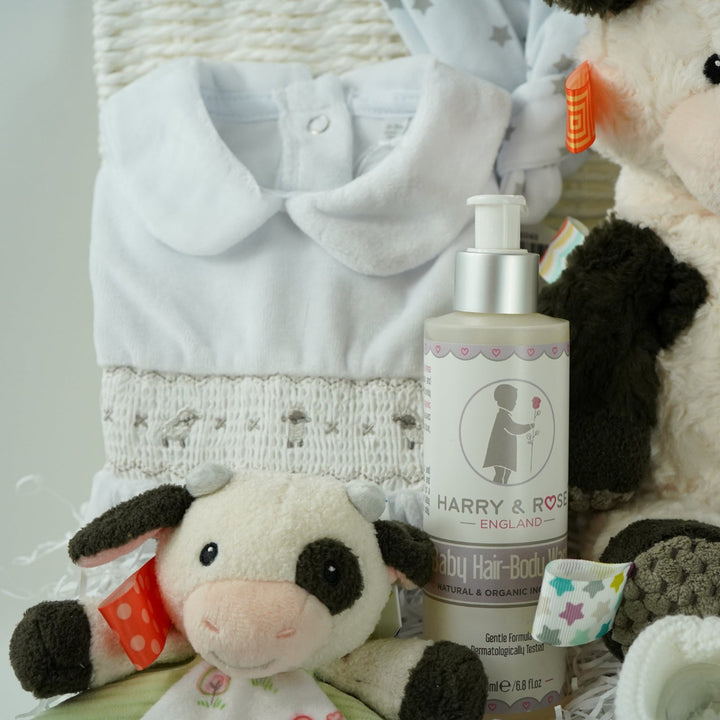 baby gift basket with a cow comforter and soft cow taggies toy, white baby sleepsuit with silver smocked sheep on bodice, organic toiletries, white knit booties and white with silver stars knot hat