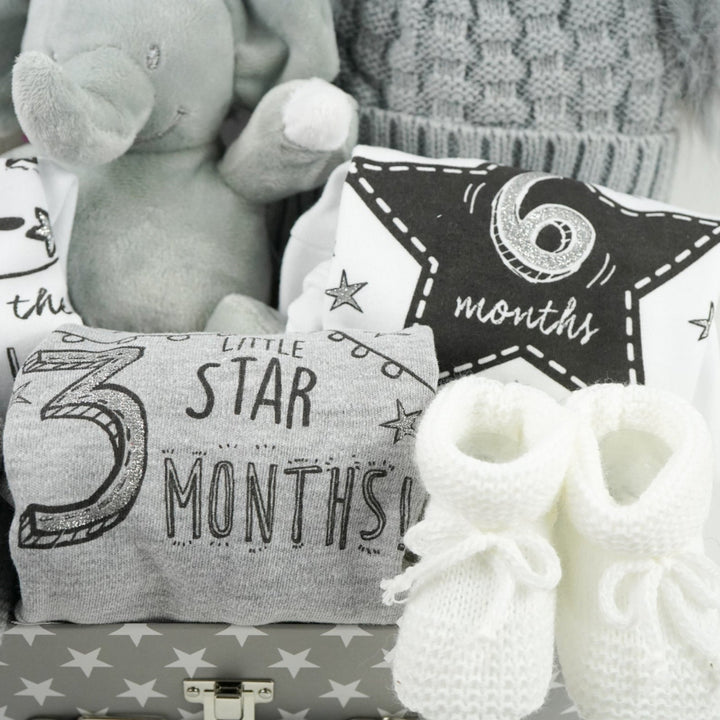 grey storage suitcase with baby items including baby hat with soft fluffy double pompoms, baby memory cards, soft elephant toy, baby set of milestone vests , white knit booties and grey knit booties