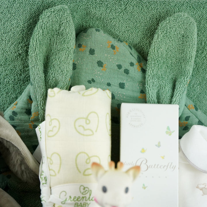 Luxury baby hamper with green hooded baby towel with ears, soft grey rabbit in green clothing set, rabbit slippers, cream and green baby swaddle, baby toiletries, green rabbit comforter, sophie la girafe teether toy, white baby sleepsuit with grey mice and magnetic fastenings all in a natural hamper basket 