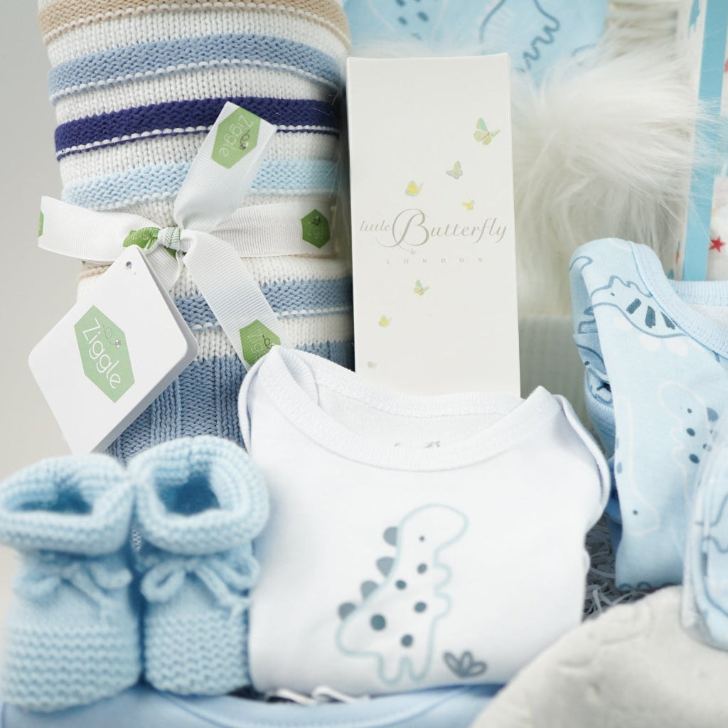 baby boy gift hamper with blue, white and cream shaped blanket, baby dinosaur clothing set, organic baby bubble bath, soft grey taggie comforter, blue booties and white double pom pom baby hat