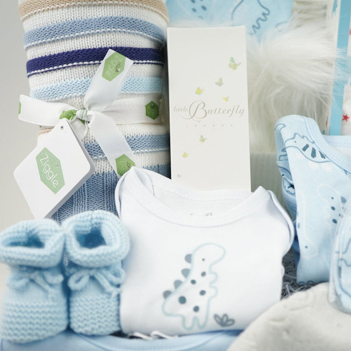 baby boy gift hamper with blue, white and cream shaped blanket, baby dinosaur clothing set, organic baby bubble bath, soft grey taggie comforter, blue booties and white double pom pom baby hat