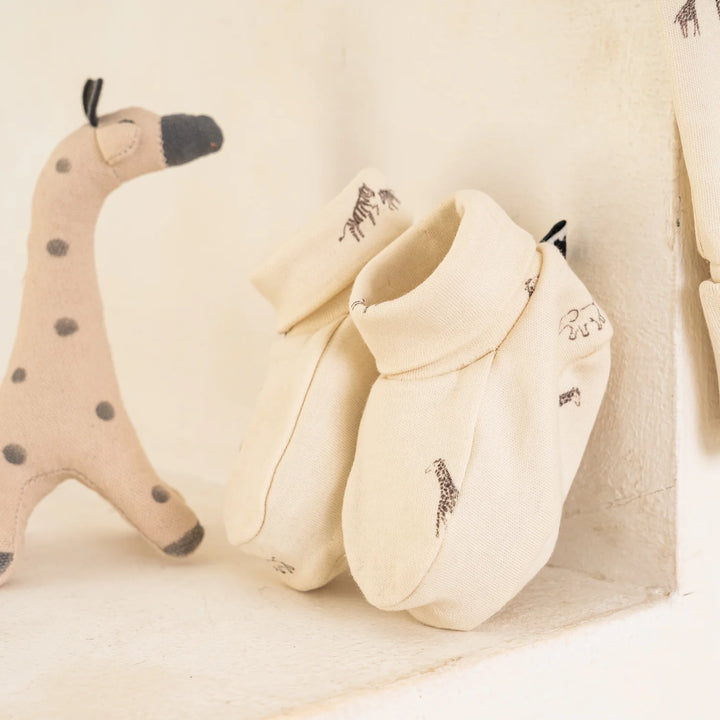 A pair of cream-colored baby booties with small embroidered giraffe motifs, displayed on a shelf next to a plush toy.