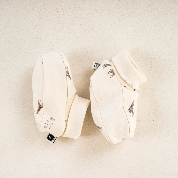 A pair of cream-colored baby booties with small black giraffe prints, laid flat on a light beige textured background.