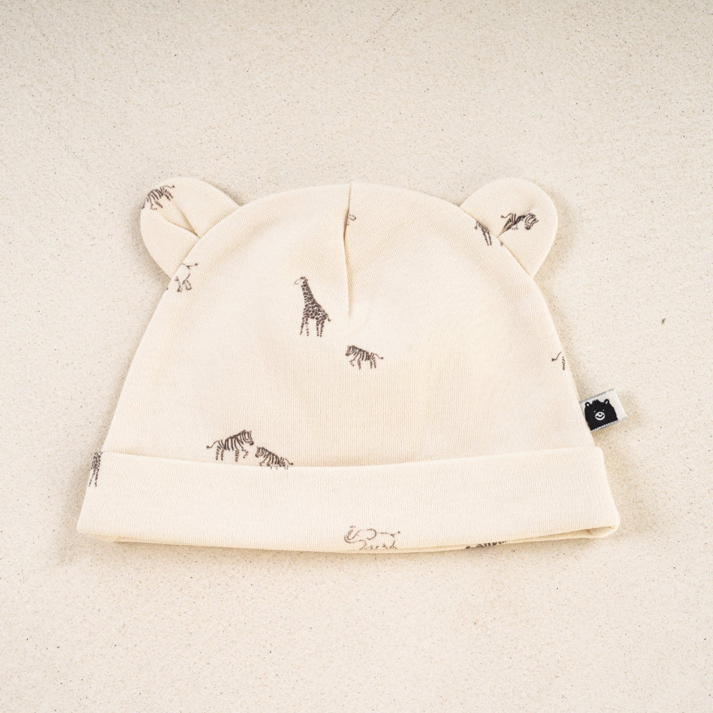 An off-white baby beanie hat with ear details and a small animal print, displayed on a textured beige background.