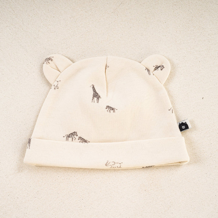 An off-white baby beanie hat with ear details and a small animal print, displayed on a textured beige background.