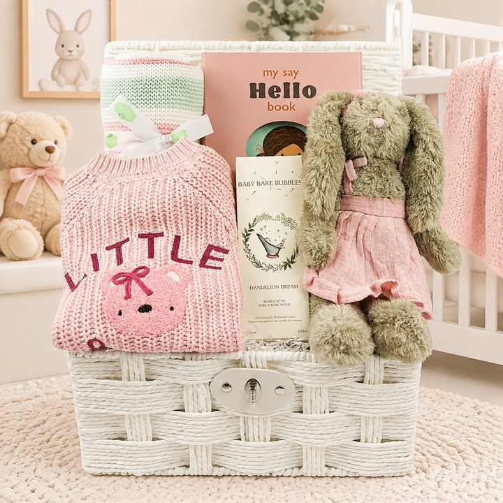A white woven baby gift basket filled with nursery-themed items, including a pink knitted baby cardigan that says 'LITTLE' BEAR' , a striped blanket, a Hello book, toiletries  and a plush green bunny in a pink dress, set in a soft nursery scene.