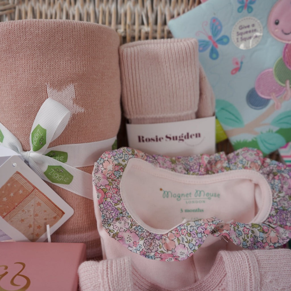 White wash hamper basket, baby gifts including baby pink cashmere cardigan, pink reversible baby blanket with stars, taggie caterpillar soft toy, taggie caterpillar crickle teether book, mum cashmere bed socks in poink, pink bump candle in a gift box, pink cashmere baby booties , pink magnetic closing baby sleepsuit with frilled collar