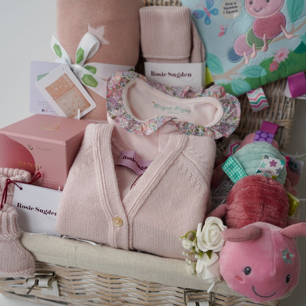 White wash hamper basket, baby gifts including baby pink cashmere cardigan, pink reversible baby blanket with stars, taggie caterpillar soft toy, taggie caterpillar crickle teether book, mum cashmere bed socks in poink, pink bump candle in a gift box, pink cashmere baby booties , pink magnetic closing baby sleepsuit with frilled collar