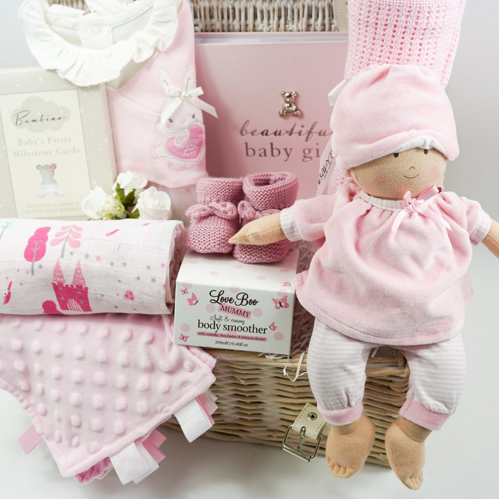 White hamper basket with Pink spanish style baby sleepsuit, baby soft doll in pink and white clothing, baby pink photo album, pink booties, baby comforter , Milestone cards , baby cellular blanket