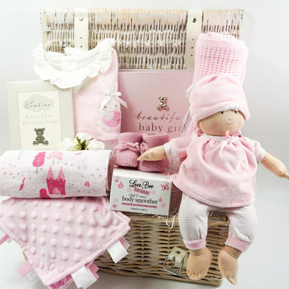White hamper basket with Pink spanish style baby sleepsuit, baby soft doll in pink and white clothing, baby pink photo album, pink booties, baby comforter , Milestone cards , baby cellular blanket