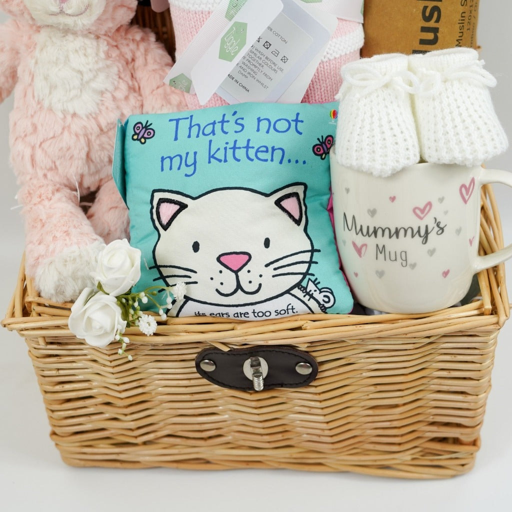 Wicker hamper with pink and white kitty soft toy, pink and white stripe heavy baby blanket, Thats Not My Kitty soft book, white baby booties, white muslin swaddle with pink flowers, mummy bone china mug