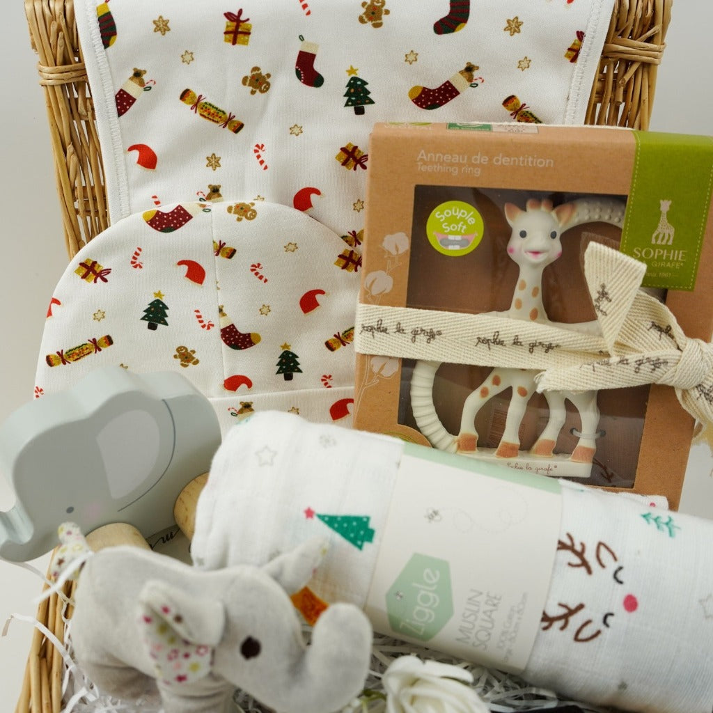 luxury cream baby christmas bib and hat set with Christmas trees, crackers, presents and stockings design, Sophie la Girafe teether, white muslin with reindeer and christmas trees, wooden push along elephant, Steiff crincle elephant rattle all in a small hamper basket